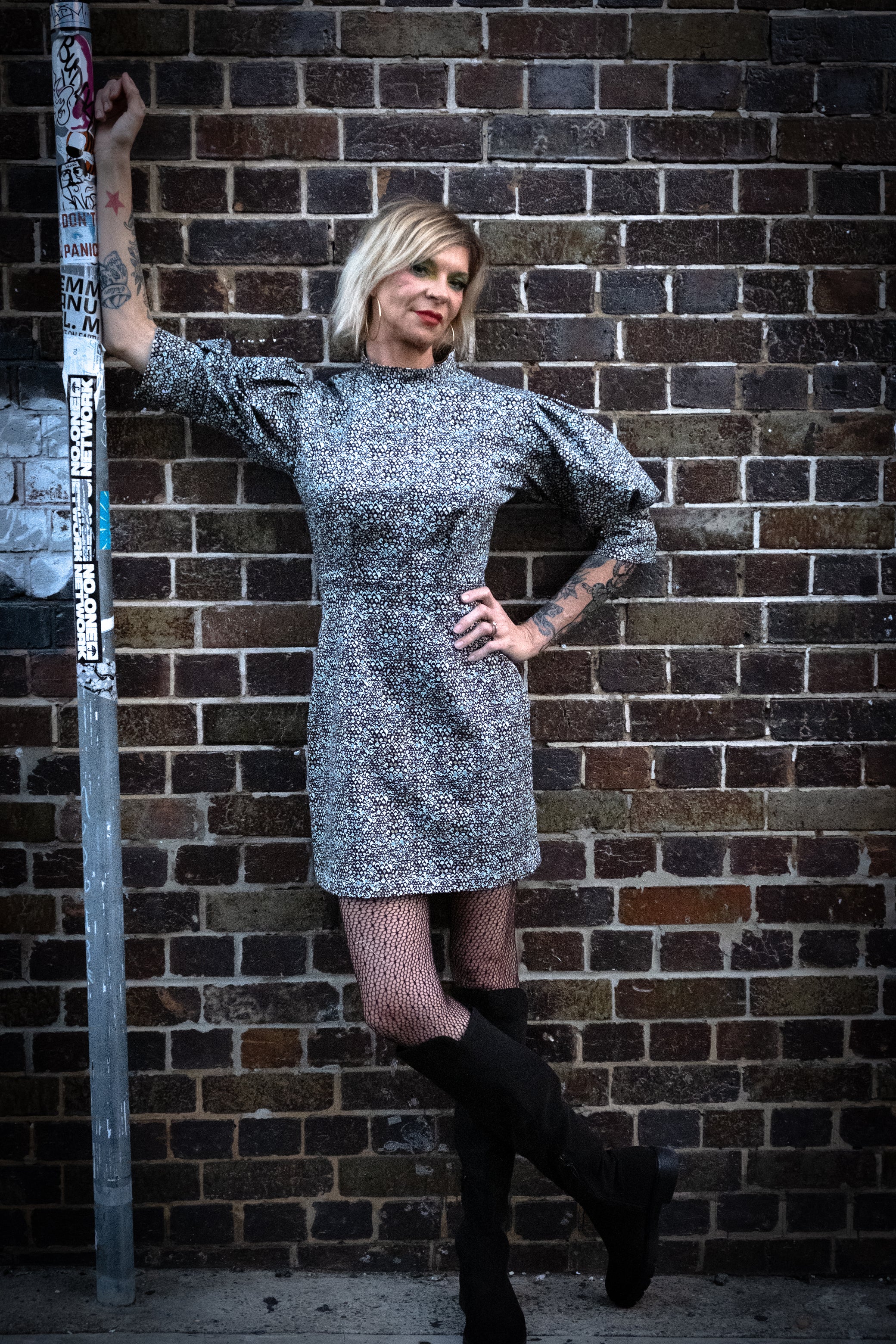 A model leaning on a pole wearing a high neck, puffy sleeve grey toned dress with black high knee boots