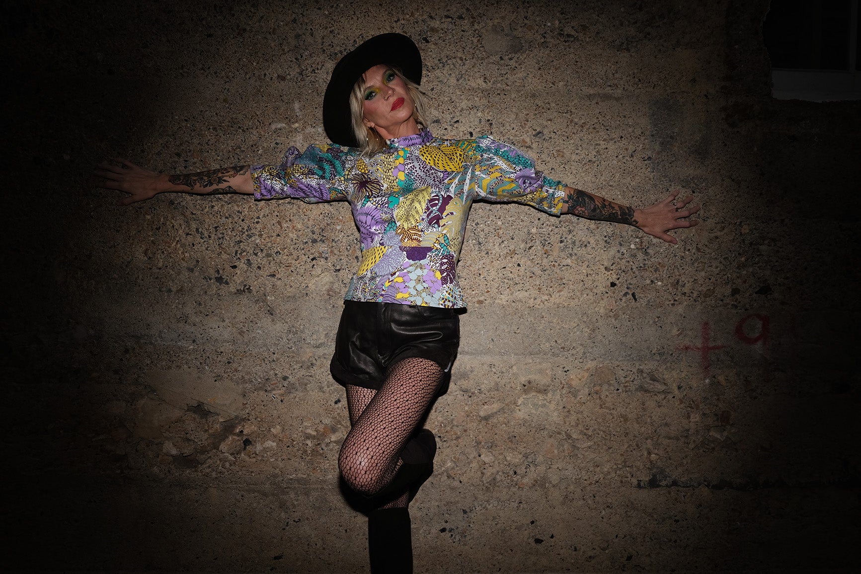 A grunge style model leaning against a wall in a colourful shirt, black shorts and a black hat.