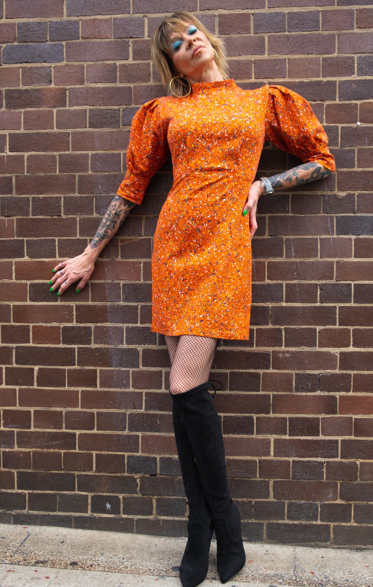 A model leaning against a brick wall wearing an orange high neck, puffy sleeves dress, and black knee-high boots. 