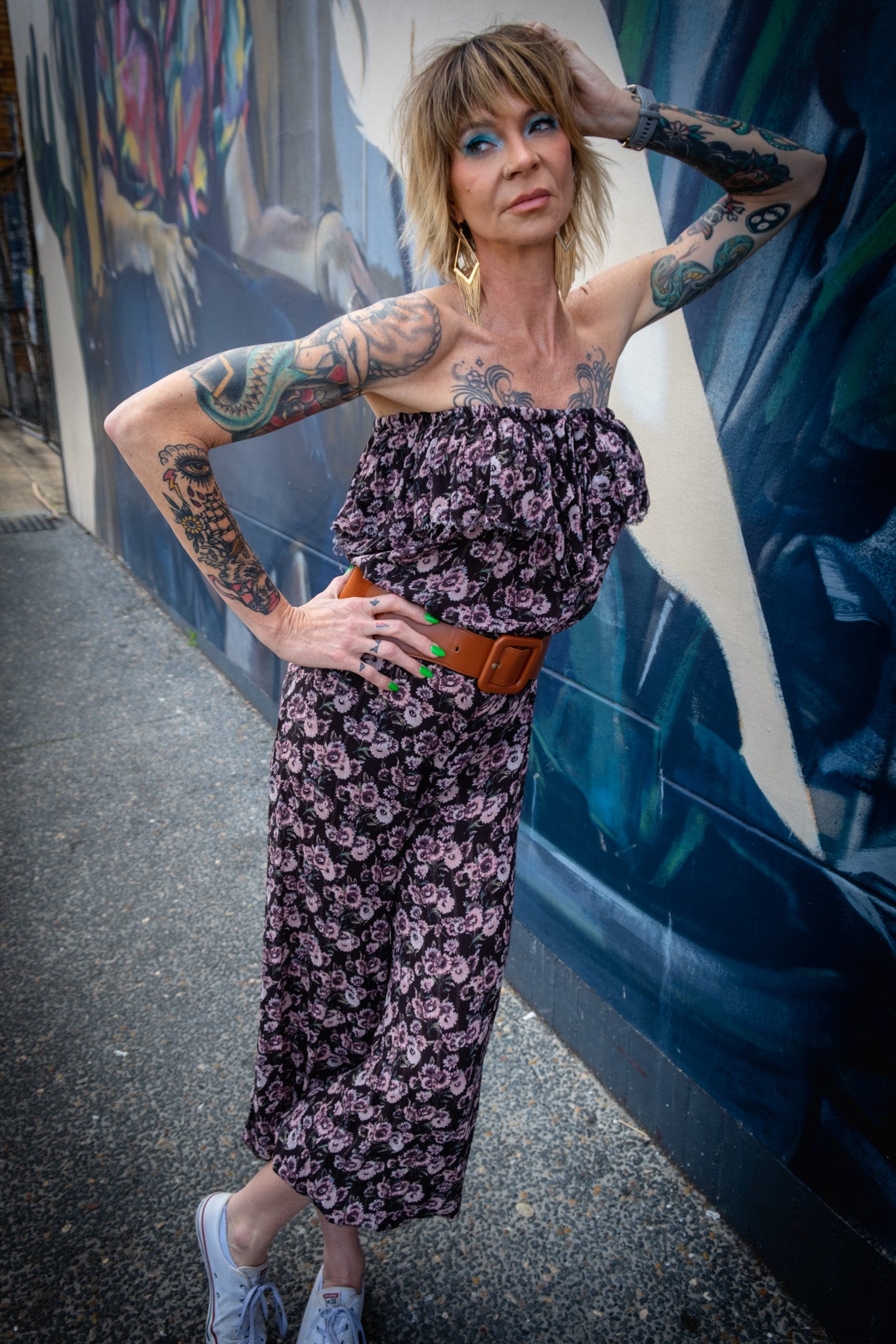 Punk style model wearing a black and purple jumpsuit with a brown belt while leaning on the wall.