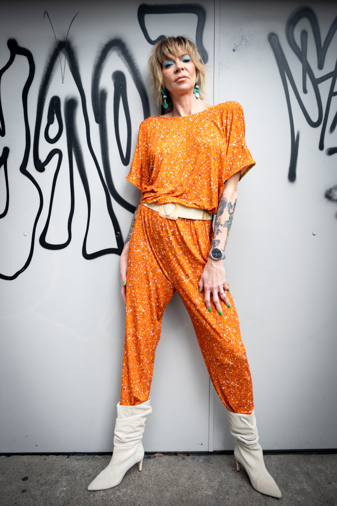 A model posing against a white wall wearing a bright orange jumpsuit, white belt, and ankle boots.
