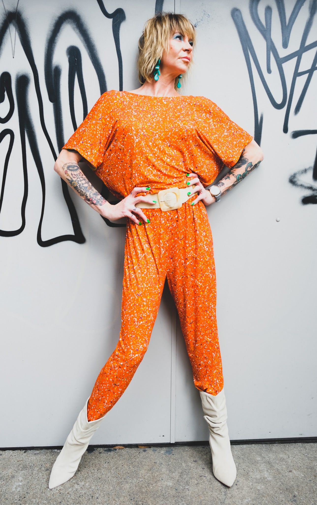 A model posing in an orange jumpsuit, white belt and ankle boots with hands on the hips