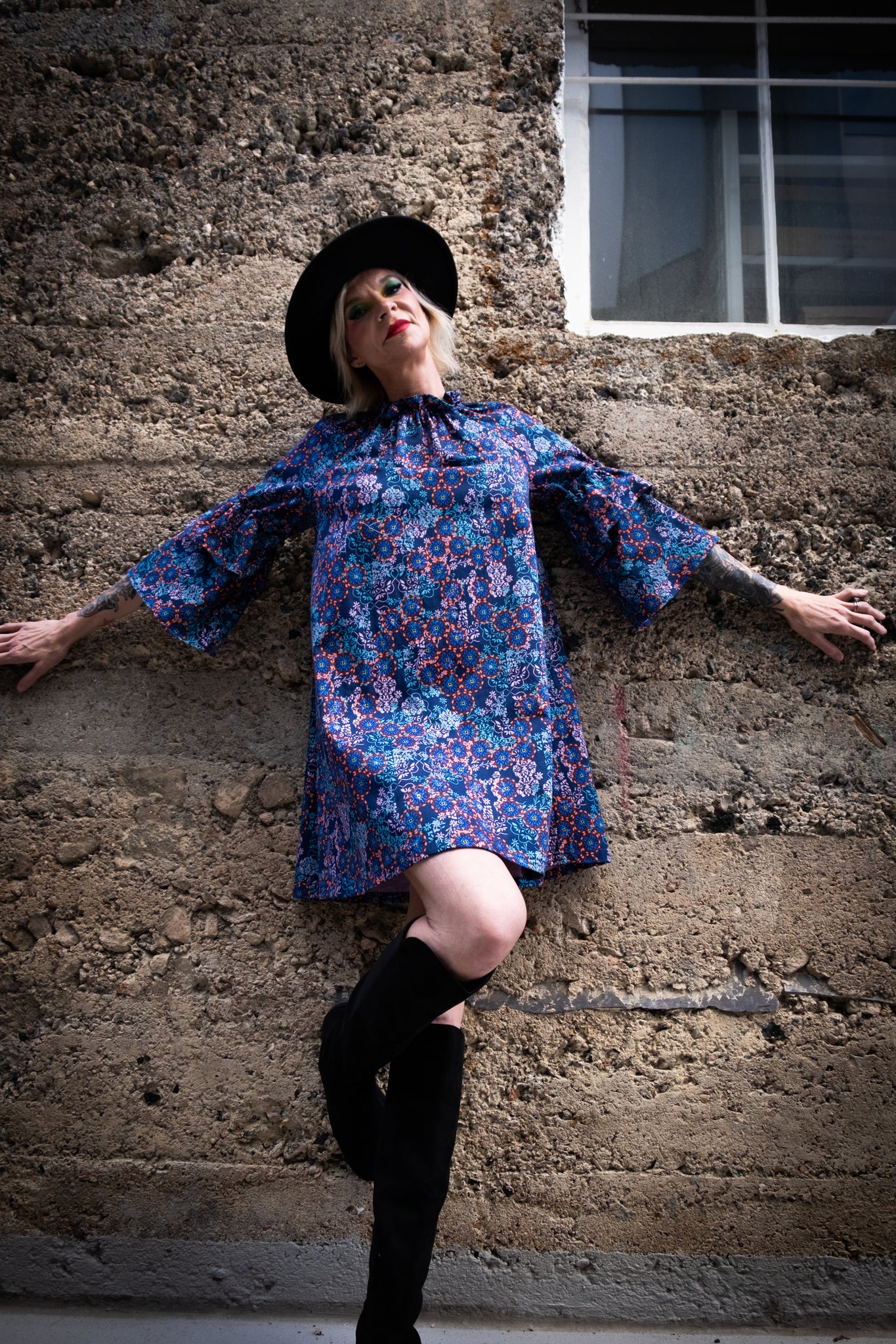 A model leaning against a wall with spread arms wearing a blue and pink dress, a black hat, and black knee-high boots. 